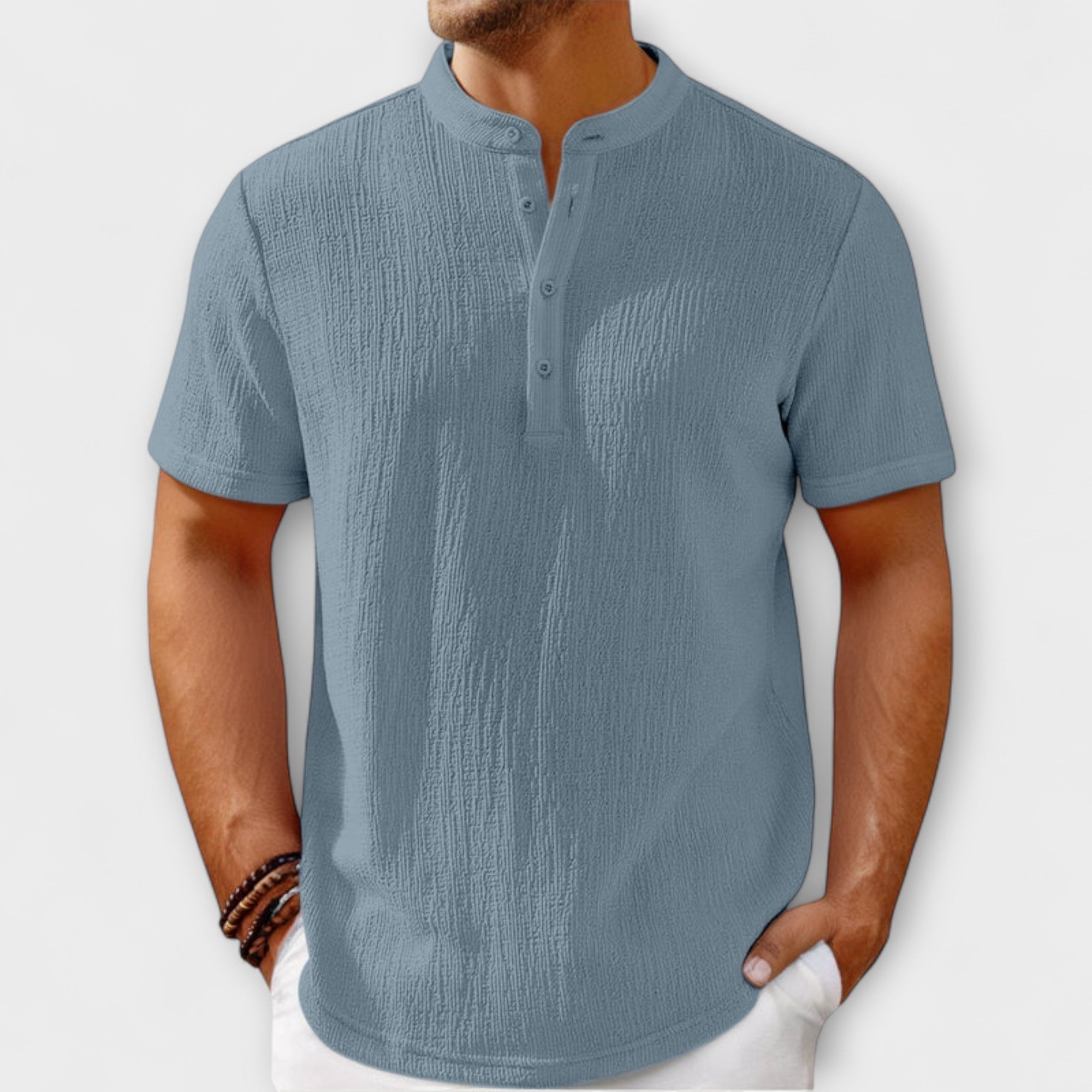 Men's short-sleeve, band-collar Henley shirt in a light cream/off-white textured linen blend fabric. The shirt has a three-button placket. The model is standing with hands in white trousers pockets, showing the relaxed fit. Focus on the linen-blend texture and the minimalist collar design.