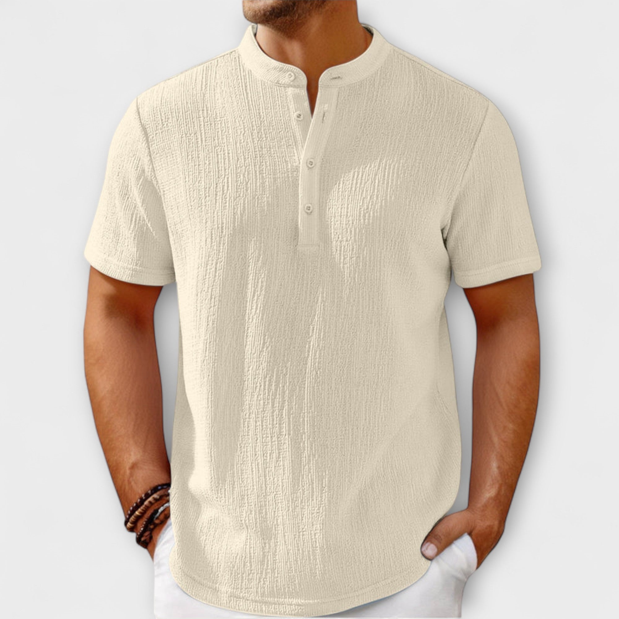 Men's short-sleeve, band-collar Henley shirt in a light cream/off-white textured linen blend fabric. The shirt has a three-button placket. The model is standing with hands in white trousers pockets, showing the relaxed fit. Focus on the linen-blend texture and the minimalist collar design.