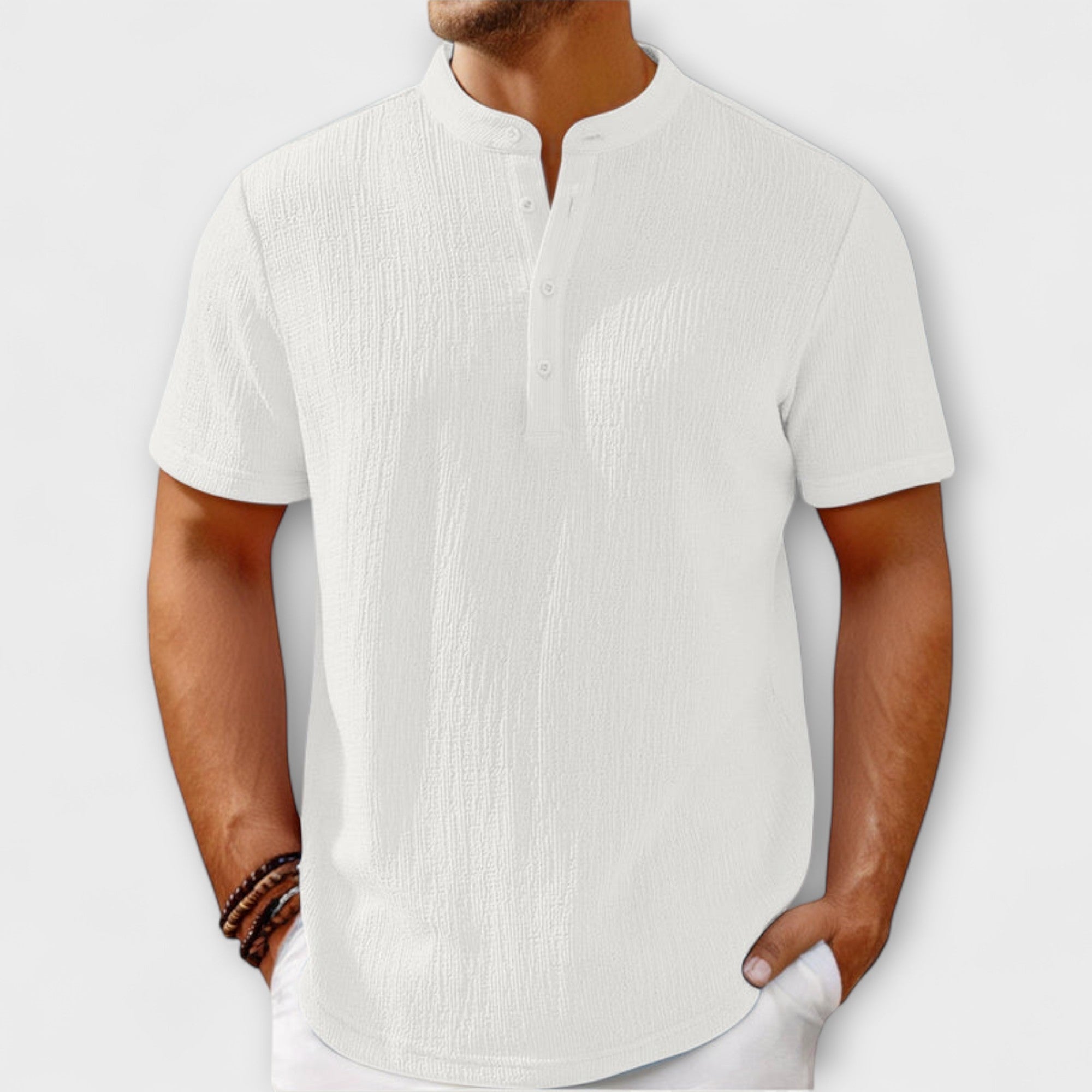 Men's short-sleeve, band-collar Henley shirt in a light cream/off-white textured linen blend fabric. The shirt has a three-button placket. The model is standing with hands in white trousers pockets, showing the relaxed fit. Focus on the linen-blend texture and the minimalist collar design.
