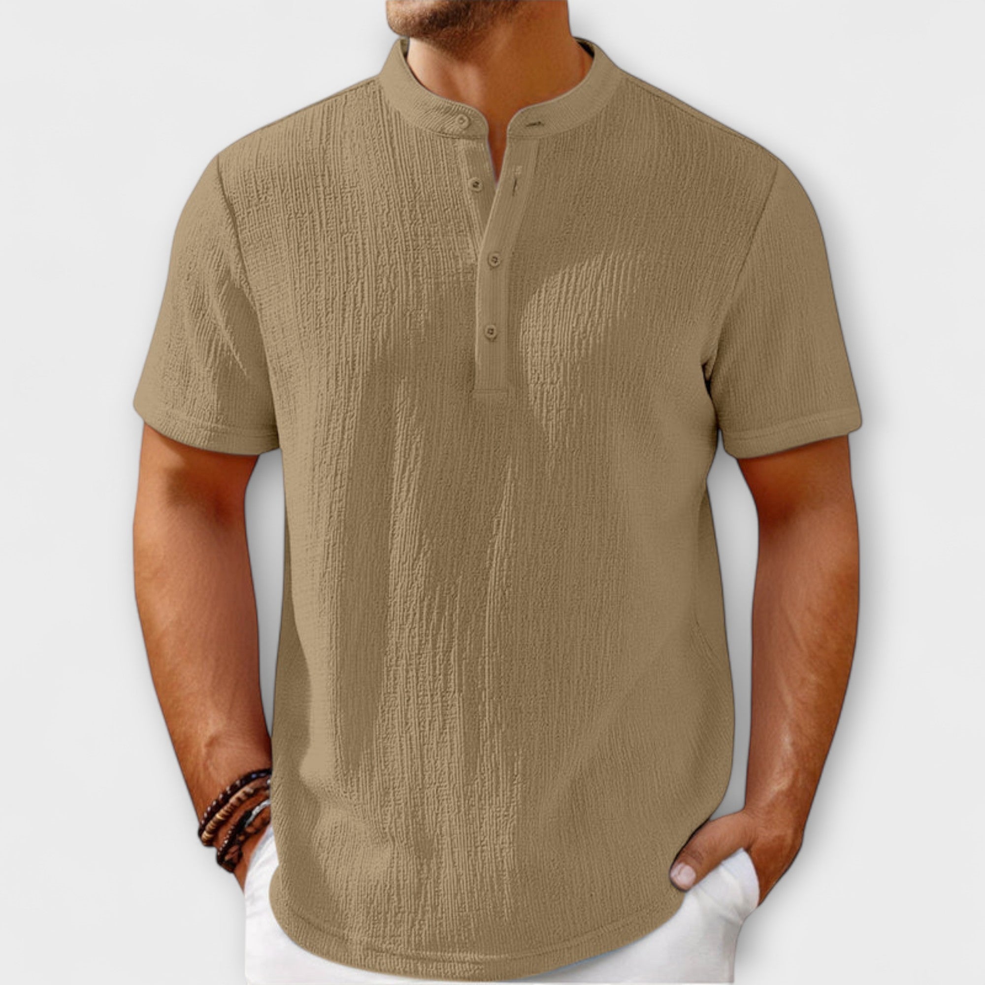Men's short-sleeve, band-collar Henley shirt in a light cream/off-white textured linen blend fabric. The shirt has a three-button placket. The model is standing with hands in white trousers pockets, showing the relaxed fit. Focus on the linen-blend texture and the minimalist collar design.