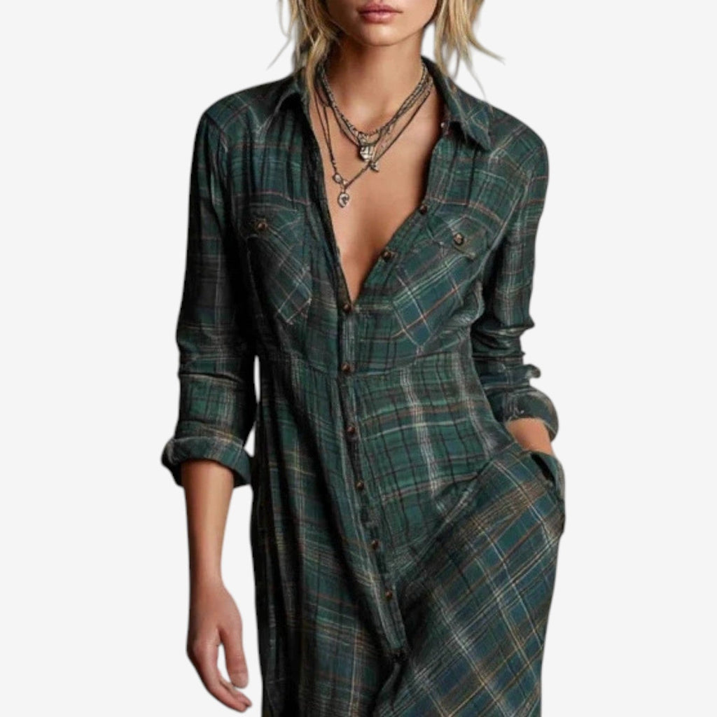 Full-length image of a woman modelling a maxi-length shirt dress in a rustic green, brown, and cream plaid/check pattern. The dress features a button-up front, a collar, and long sleeves that are rolled up. A matching fabric belt is tied around the waist, and the skirt flows to the ankles. Focus on the fabric pattern, the dress's length, and the waist-cinching detail.