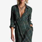 Full-length image of a woman modelling a maxi-length shirt dress in a rustic green, brown, and cream plaid/check pattern. The dress features a button-up front, a collar, and long sleeves that are rolled up. A matching fabric belt is tied around the waist, and the skirt flows to the ankles. Focus on the fabric pattern, the dress's length, and the waist-cinching detail.