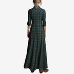 Full-length image of a woman modelling a maxi-length shirt dress in a rustic green, brown, and cream plaid/check pattern. The dress features a button-up front, a collar, and long sleeves that are rolled up. A matching fabric belt is tied around the waist, and the skirt flows to the ankles. Focus on the fabric pattern, the dress's length, and the waist-cinching detail.