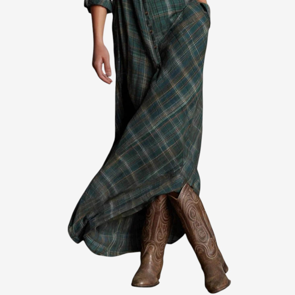 Full-length image of a woman modelling a maxi-length shirt dress in a rustic green, brown, and cream plaid/check pattern. The dress features a button-up front, a collar, and long sleeves that are rolled up. A matching fabric belt is tied around the waist, and the skirt flows to the ankles. Focus on the fabric pattern, the dress's length, and the waist-cinching detail.