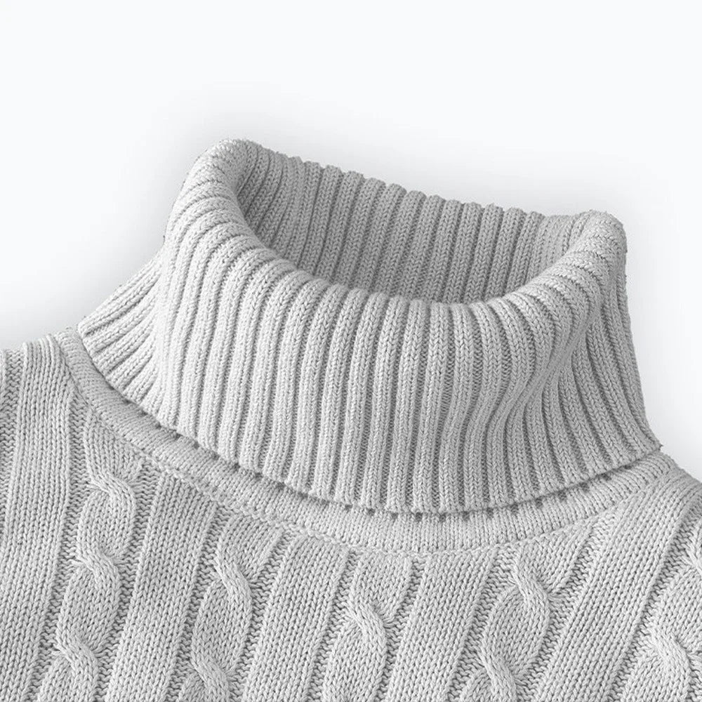 Folded men's turtleneck/roll-neck jumper in a light beige/oatmeal colour. The knit features a subtle vertical cable pattern down the body. The turtleneck and cuffs are ribbed. The sweater is styled next to a black leather men's wristwatch. Focus on the fine cable texture and the clean fold.