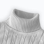 Folded men's turtleneck/roll-neck jumper in a light beige/oatmeal colour. The knit features a subtle vertical cable pattern down the body. The turtleneck and cuffs are ribbed. The sweater is styled next to a black leather men's wristwatch. Focus on the fine cable texture and the clean fold.