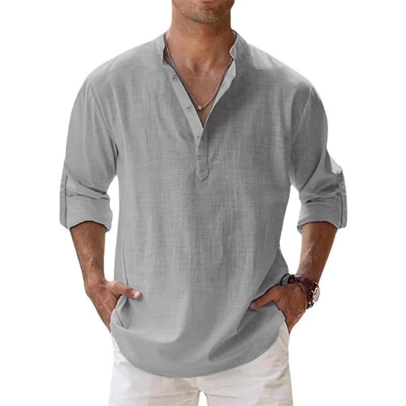 Men's long-sleeve popover shirt in white, featuring a Grandad/band collar and a short buttoned Henley placket. The shirt is made from a light, textured linen-blend fabric. The model is standing with hands in pockets, showcasing the relaxed, slightly oversized fit and rolled-up sleeves. Focus on the texture and the minimalist collar.