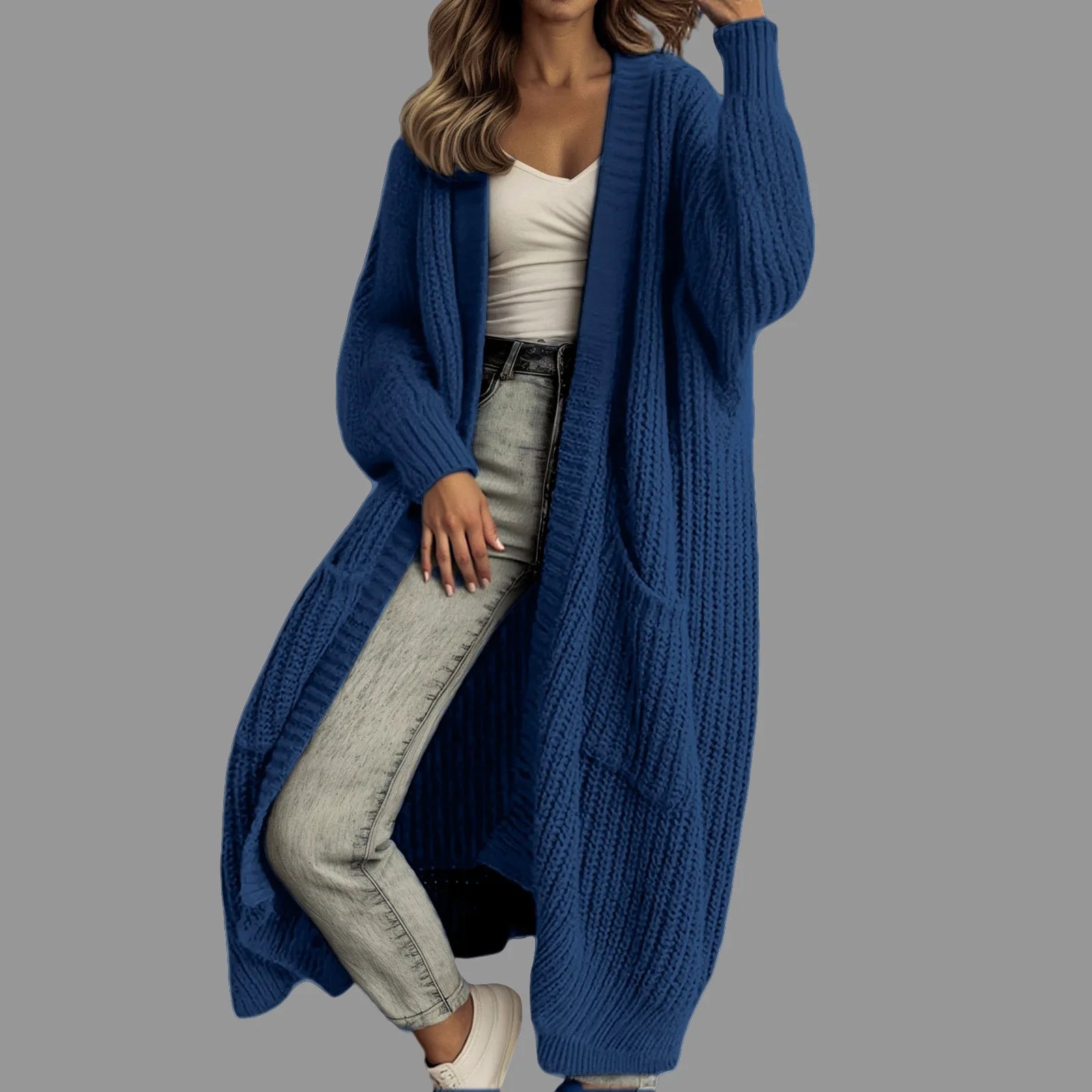 Full-length view of a women's light beige/oatmeal chunky ribbed knit duster/longline cardigan. The cardigan has a relaxed, open-front fit, dropped shoulders, and two large patch pockets. It is styled casually over a white top and blue denim jeans. Focus on the vertical rib texture and the garment's length.