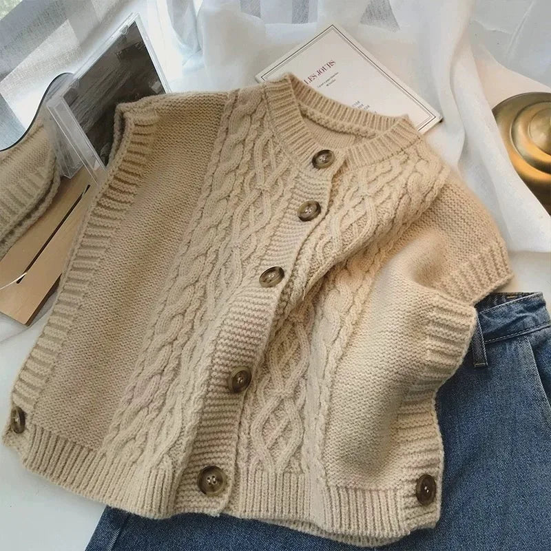 Close-up of a women's beige/cream cable knit button-up gilet/vest worn over a white long-sleeve shirt and paired with light wash denim jeans. The gilet features chunky, prominent cable knit patterns and large brown tortoiseshell buttons down the front. Focus on the texture and layering.