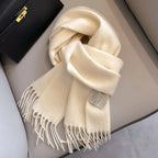Close-up of a folded, creamy white/ivory cashmere-feel scarf draped on a chair next to a black leather handbag. The scarf features a soft, brushed texture and neat white knotted fringe tassels at the ends. Focus on the luxurious texture and the fringe detail.