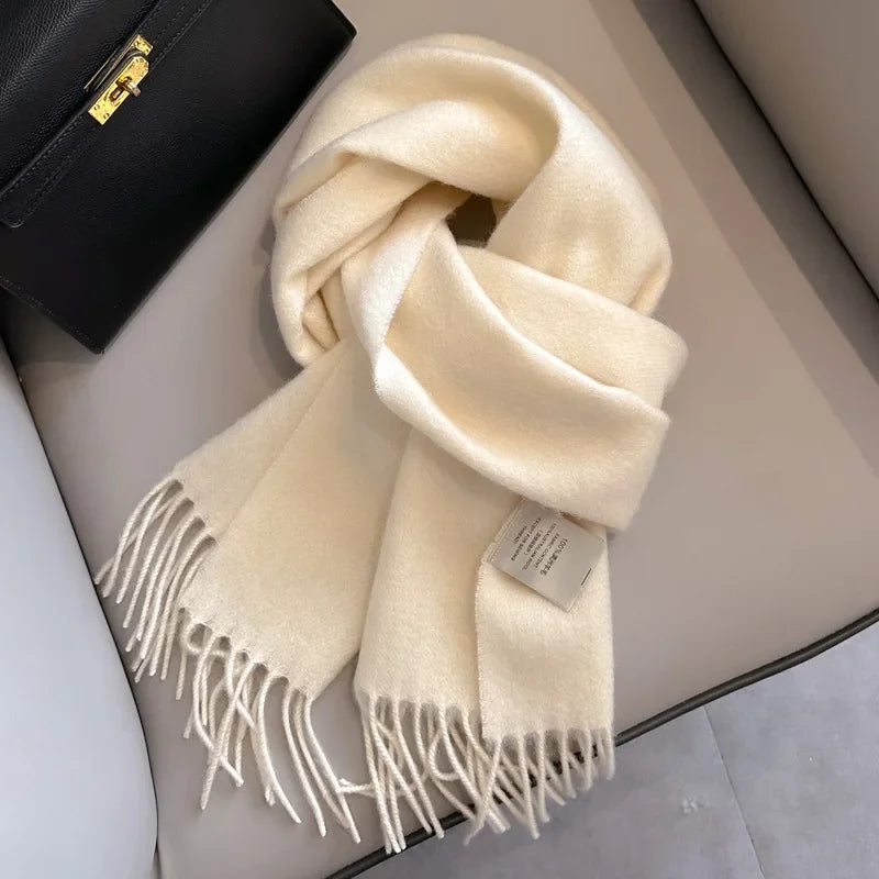 Close-up of a folded, creamy white/ivory cashmere-feel scarf draped on a chair next to a black leather handbag. The scarf features a soft, brushed texture and neat white knotted fringe tassels at the ends. Focus on the luxurious texture and the fringe detail.