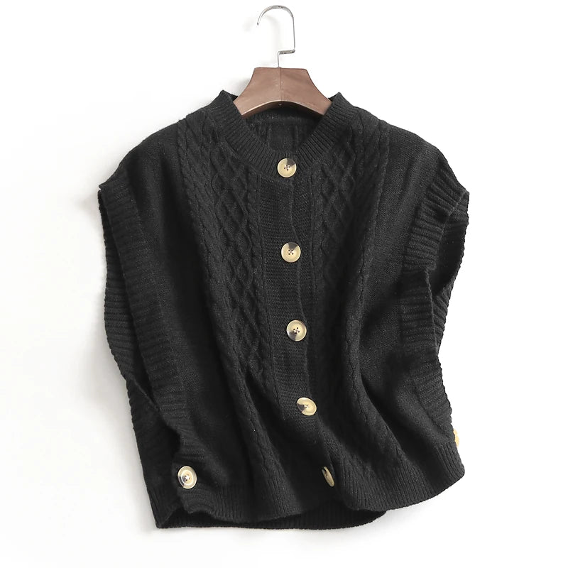 Close-up of a women's beige/cream cable knit button-up gilet/vest worn over a white long-sleeve shirt and paired with light wash denim jeans. The gilet features chunky, prominent cable knit patterns and large brown tortoiseshell buttons down the front. Focus on the texture and layering.