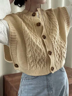 Close-up of a women's beige/cream cable knit button-up gilet/vest worn over a white long-sleeve shirt and paired with light wash denim jeans. The gilet features chunky, prominent cable knit patterns and large brown tortoiseshell buttons down the front. Focus on the texture and layering.