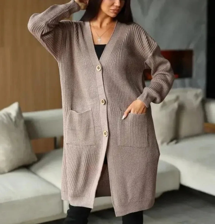 Full-length view of a women's light beige/oatmeal chunky ribbed knit duster/longline cardigan. The cardigan has a relaxed, open-front fit, dropped shoulders, and two large patch pockets. It is styled casually over a white top and blue denim jeans. Focus on the vertical rib texture and the garment's length.