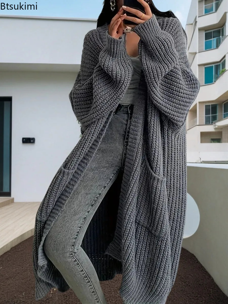 Full-length view of a women's light beige/oatmeal chunky ribbed knit duster/longline cardigan. The cardigan has a relaxed, open-front fit, dropped shoulders, and two large patch pockets. It is styled casually over a white top and blue denim jeans. Focus on the vertical rib texture and the garment's length.