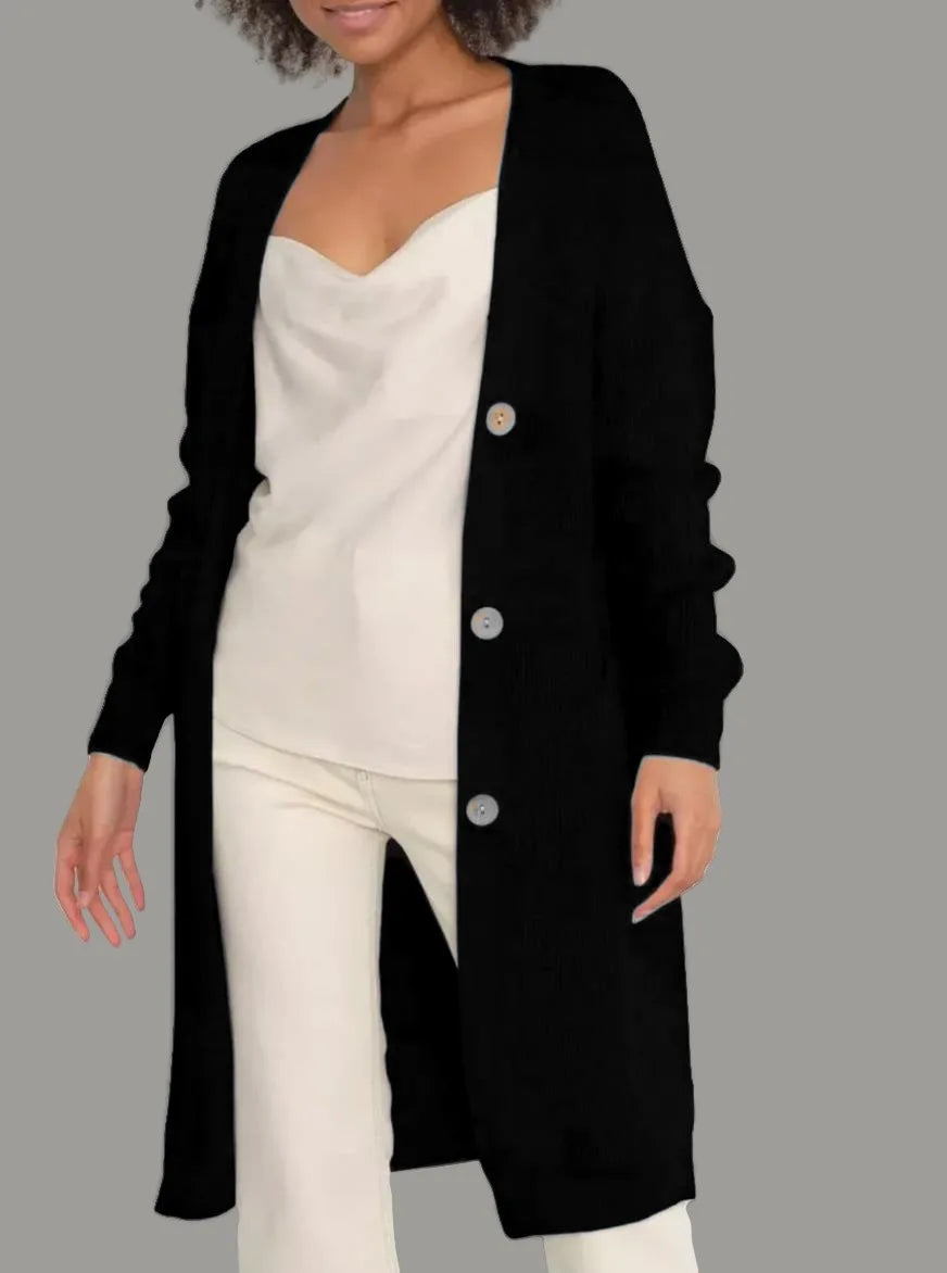 Full-length view of a women's light beige/oatmeal chunky ribbed knit duster/longline cardigan. The cardigan has a relaxed, open-front fit, dropped shoulders, and two large patch pockets. It is styled casually over a white top and blue denim jeans. Focus on the vertical rib texture and the garment's length.