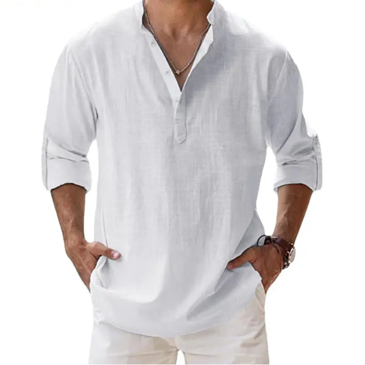 Men's long-sleeve popover shirt in white, featuring a Grandad/band collar and a short buttoned Henley placket. The shirt is made from a light, textured linen-blend fabric. The model is standing with hands in pockets, showcasing the relaxed, slightly oversized fit and rolled-up sleeves. Focus on the texture and the minimalist collar.