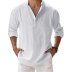 Men's long-sleeve popover shirt in white, featuring a Grandad/band collar and a short buttoned Henley placket. The shirt is made from a light, textured linen-blend fabric. The model is standing with hands in pockets, showcasing the relaxed, slightly oversized fit and rolled-up sleeves. Focus on the texture and the minimalist collar.
