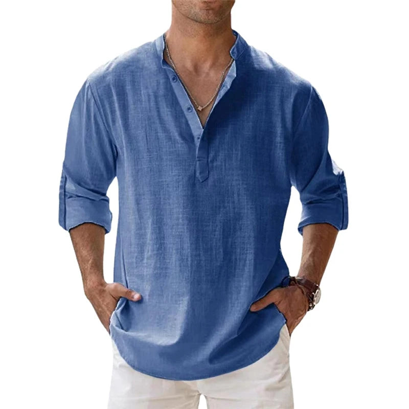 Men's long-sleeve popover shirt in white, featuring a Grandad/band collar and a short buttoned Henley placket. The shirt is made from a light, textured linen-blend fabric. The model is standing with hands in pockets, showcasing the relaxed, slightly oversized fit and rolled-up sleeves. Focus on the texture and the minimalist collar.