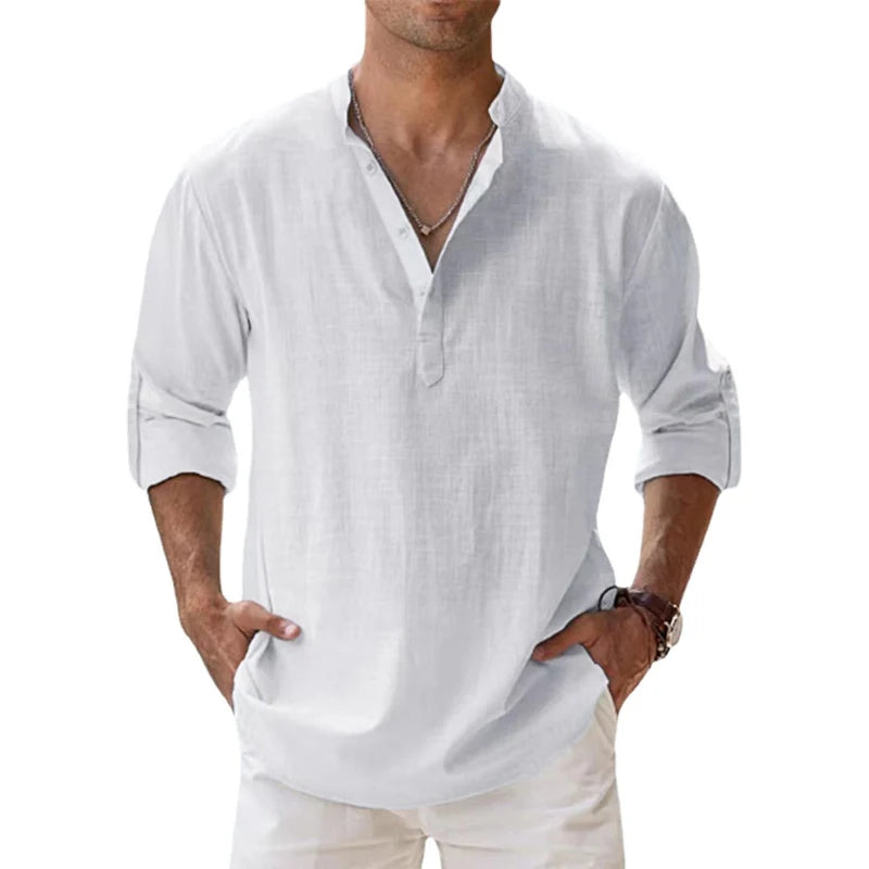 Men's long-sleeve popover shirt in white, featuring a Grandad/band collar and a short buttoned Henley placket. The shirt is made from a light, textured linen-blend fabric. The model is standing with hands in pockets, showcasing the relaxed, slightly oversized fit and rolled-up sleeves. Focus on the texture and the minimalist collar.