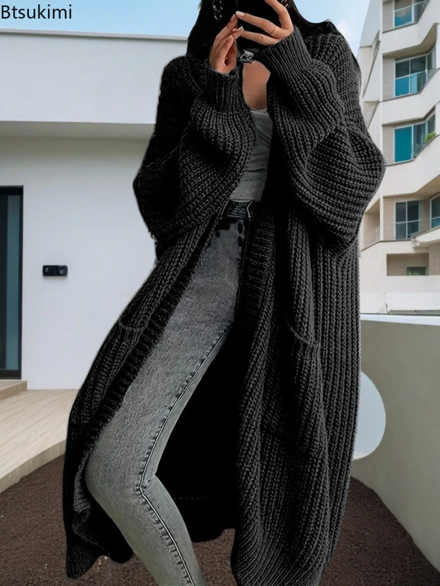 Full-length view of a women's light beige/oatmeal chunky ribbed knit duster/longline cardigan. The cardigan has a relaxed, open-front fit, dropped shoulders, and two large patch pockets. It is styled casually over a white top and blue denim jeans. Focus on the vertical rib texture and the garment's length.