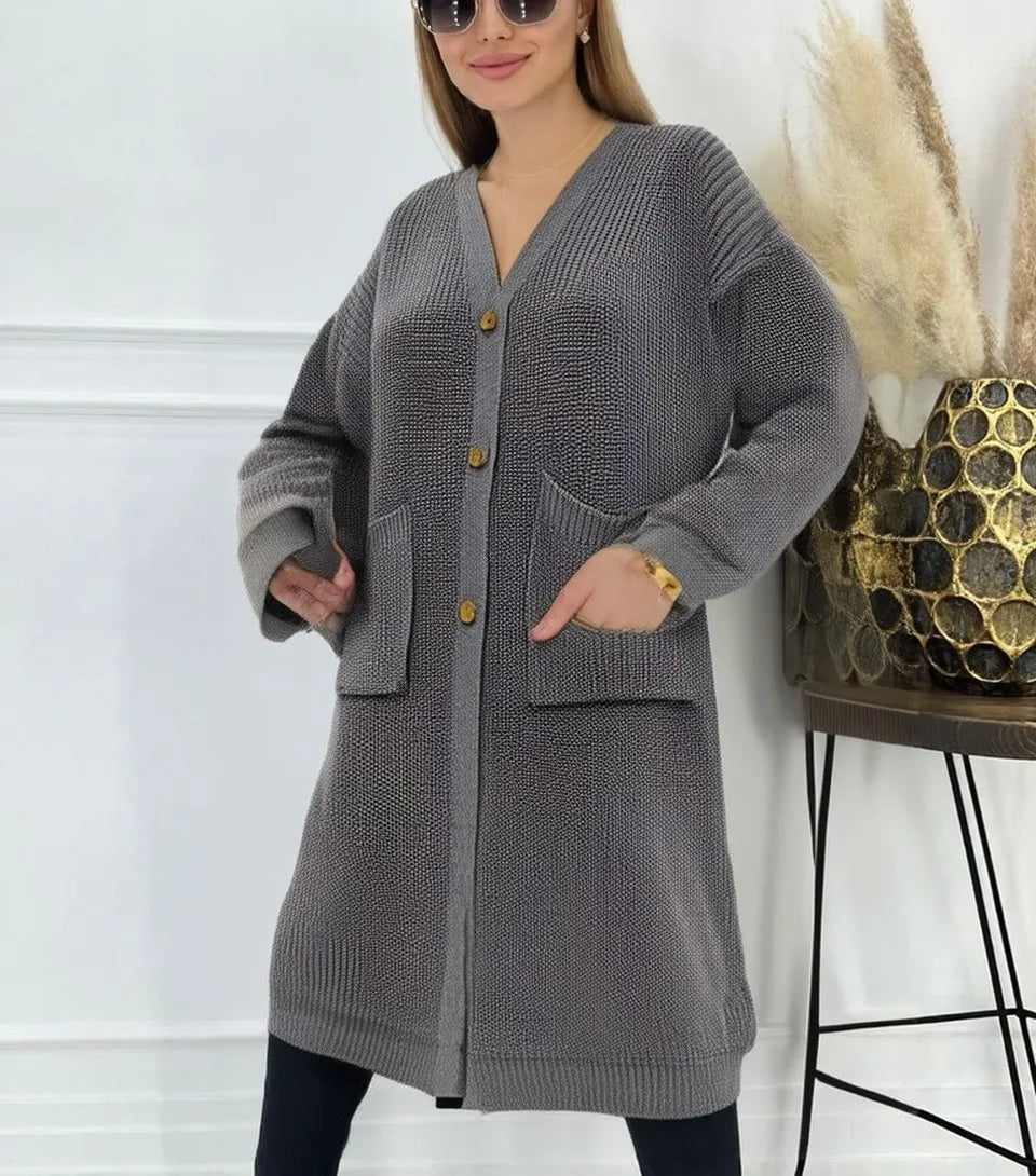 Full-length view of a women's light beige/oatmeal chunky ribbed knit duster/longline cardigan. The cardigan has a relaxed, open-front fit, dropped shoulders, and two large patch pockets. It is styled casually over a white top and blue denim jeans. Focus on the vertical rib texture and the garment's length.