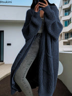 Full-length view of a women's light beige/oatmeal chunky ribbed knit duster/longline cardigan. The cardigan has a relaxed, open-front fit, dropped shoulders, and two large patch pockets. It is styled casually over a white top and blue denim jeans. Focus on the vertical rib texture and the garment's length.