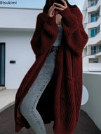 Full-length view of a women's light beige/oatmeal chunky ribbed knit duster/longline cardigan. The cardigan has a relaxed, open-front fit, dropped shoulders, and two large patch pockets. It is styled casually over a white top and blue denim jeans. Focus on the vertical rib texture and the garment's length.