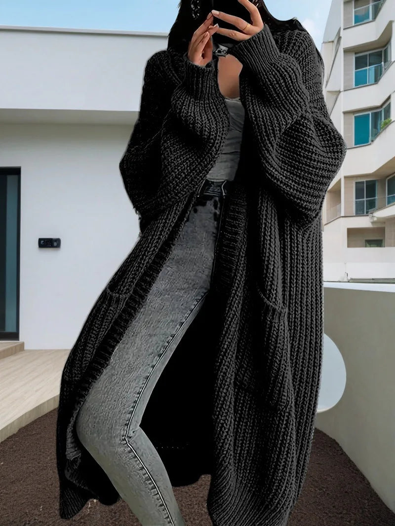 Full-length view of a women's light beige/oatmeal chunky ribbed knit duster/longline cardigan. The cardigan has a relaxed, open-front fit, dropped shoulders, and two large patch pockets. It is styled casually over a white top and blue denim jeans. Focus on the vertical rib texture and the garment's length.