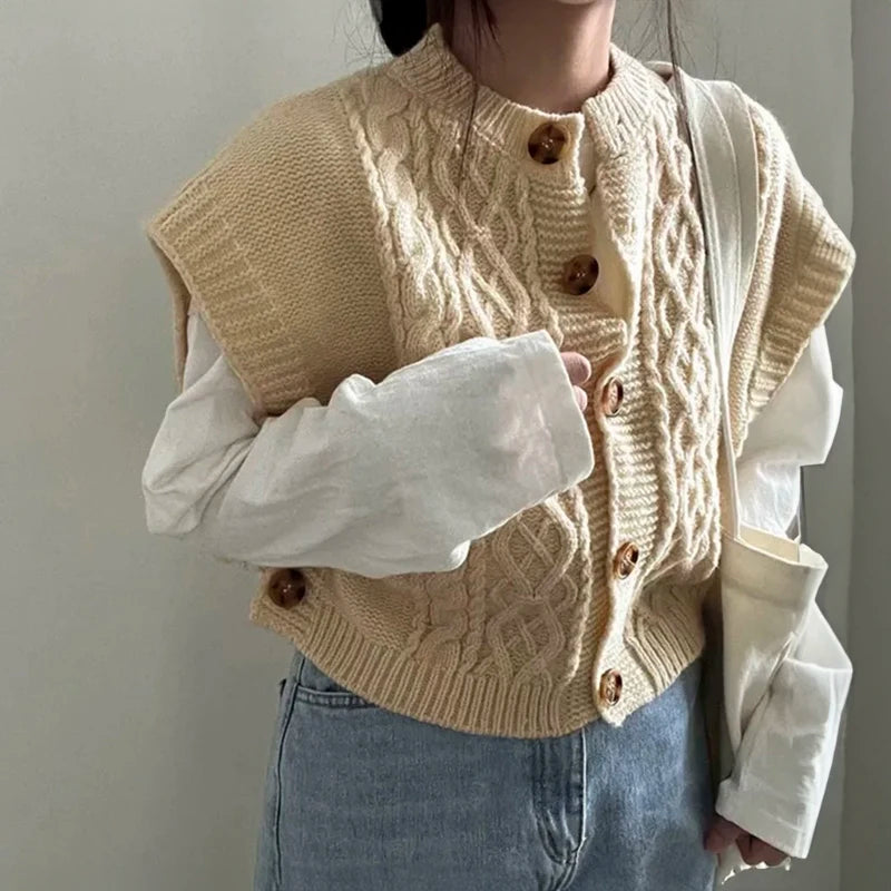 Close-up of a women's beige/cream cable knit button-up gilet/vest worn over a white long-sleeve shirt and paired with light wash denim jeans. The gilet features chunky, prominent cable knit patterns and large brown tortoiseshell buttons down the front. Focus on the texture and layering.