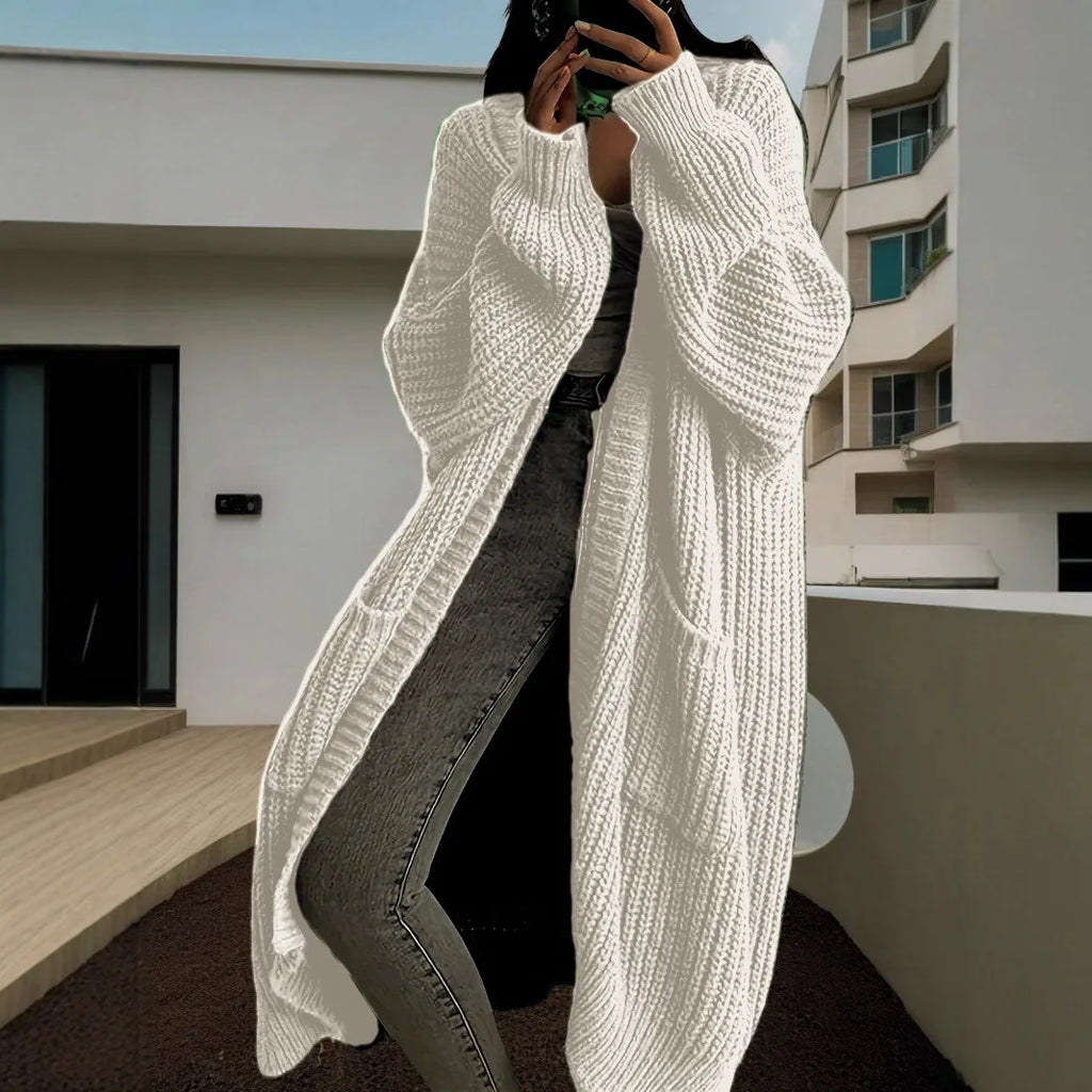 Full-length view of a women's light beige/oatmeal chunky ribbed knit duster/longline cardigan. The cardigan has a relaxed, open-front fit, dropped shoulders, and two large patch pockets. It is styled casually over a white top and blue denim jeans. Focus on the vertical rib texture and the garment's length.
