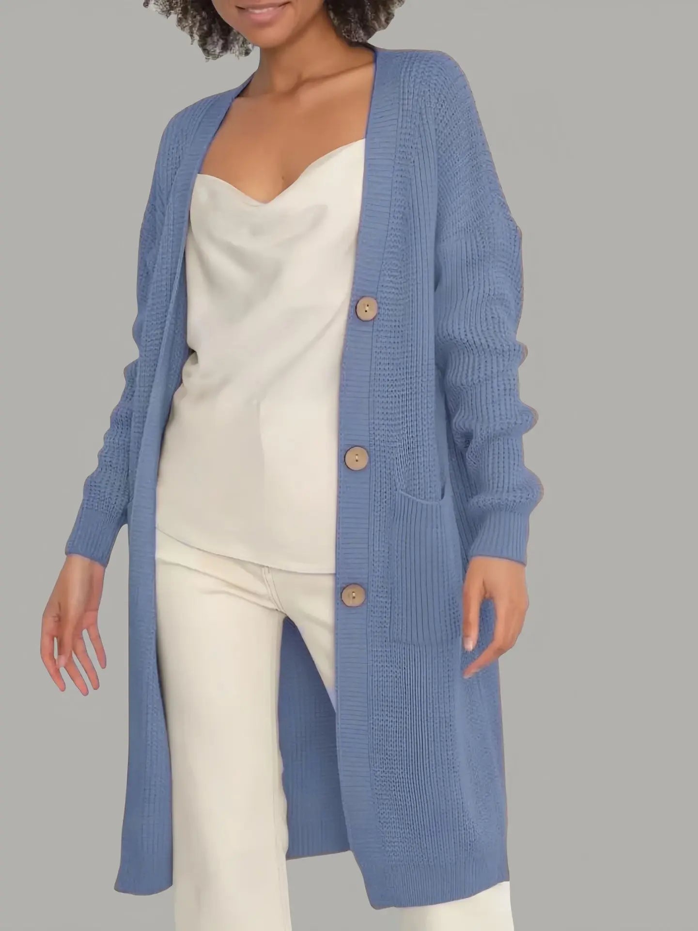 Full-length view of a women's light beige/oatmeal chunky ribbed knit duster/longline cardigan. The cardigan has a relaxed, open-front fit, dropped shoulders, and two large patch pockets. It is styled casually over a white top and blue denim jeans. Focus on the vertical rib texture and the garment's length.