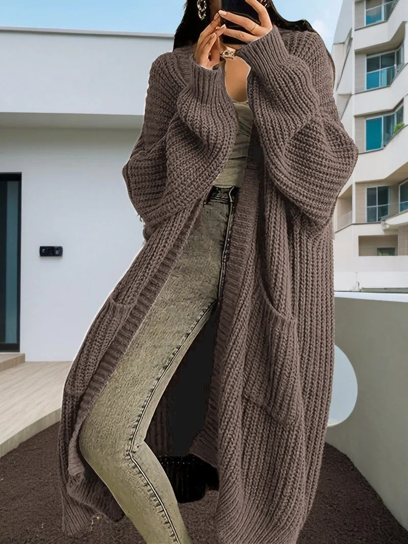 Full-length view of a women's light beige/oatmeal chunky ribbed knit duster/longline cardigan. The cardigan has a relaxed, open-front fit, dropped shoulders, and two large patch pockets. It is styled casually over a white top and blue denim jeans. Focus on the vertical rib texture and the garment's length.