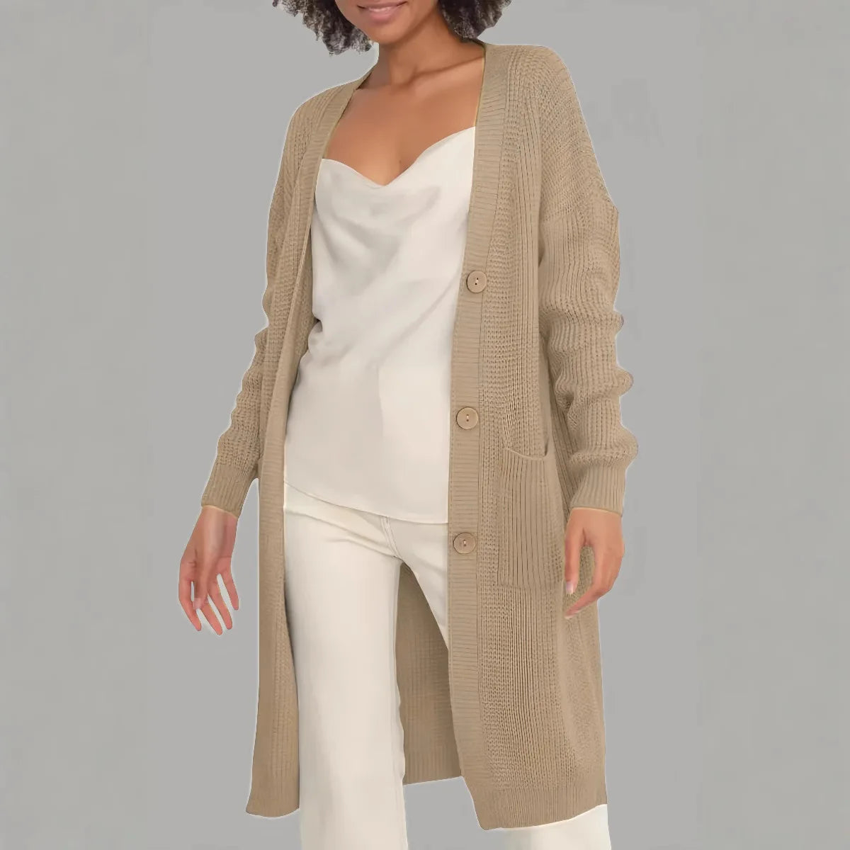 Full-length view of a women's light beige/oatmeal chunky ribbed knit duster/longline cardigan. The cardigan has a relaxed, open-front fit, dropped shoulders, and two large patch pockets. It is styled casually over a white top and blue denim jeans. Focus on the vertical rib texture and the garment's length.