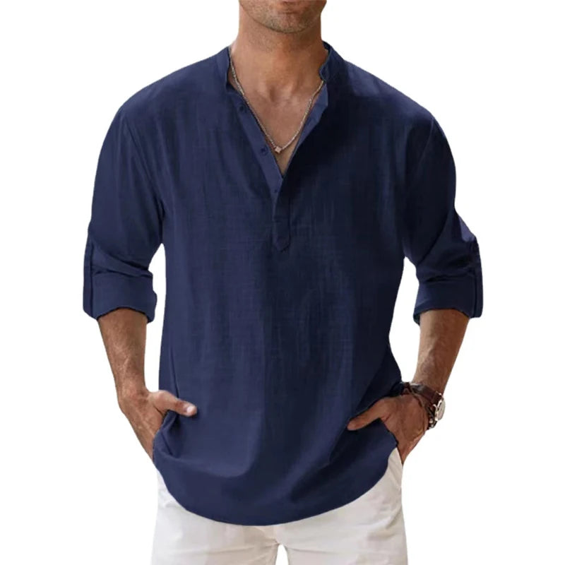 Men's long-sleeve popover shirt in white, featuring a Grandad/band collar and a short buttoned Henley placket. The shirt is made from a light, textured linen-blend fabric. The model is standing with hands in pockets, showcasing the relaxed, slightly oversized fit and rolled-up sleeves. Focus on the texture and the minimalist collar.