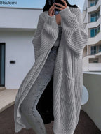 Full-length view of a women's light beige/oatmeal chunky ribbed knit duster/longline cardigan. The cardigan has a relaxed, open-front fit, dropped shoulders, and two large patch pockets. It is styled casually over a white top and blue denim jeans. Focus on the vertical rib texture and the garment's length.