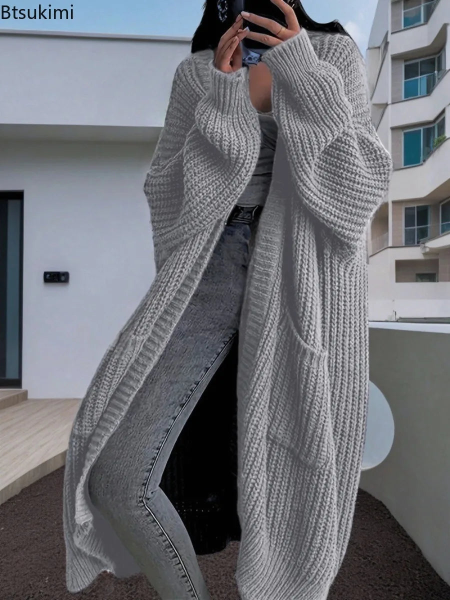 Full-length view of a women's light beige/oatmeal chunky ribbed knit duster/longline cardigan. The cardigan has a relaxed, open-front fit, dropped shoulders, and two large patch pockets. It is styled casually over a white top and blue denim jeans. Focus on the vertical rib texture and the garment's length.
