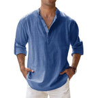 Men's long-sleeve popover shirt in white, featuring a Grandad/band collar and a short buttoned Henley placket. The shirt is made from a light, textured linen-blend fabric. The model is standing with hands in pockets, showcasing the relaxed, slightly oversized fit and rolled-up sleeves. Focus on the texture and the minimalist collar.