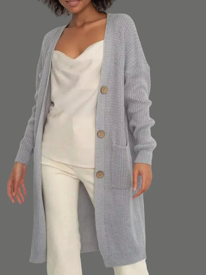 Full-length view of a women's light beige/oatmeal chunky ribbed knit duster/longline cardigan. The cardigan has a relaxed, open-front fit, dropped shoulders, and two large patch pockets. It is styled casually over a white top and blue denim jeans. Focus on the vertical rib texture and the garment's length.