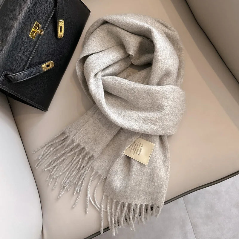 Close-up of a folded, creamy white/ivory cashmere-feel scarf draped on a chair next to a black leather handbag. The scarf features a soft, brushed texture and neat white knotted fringe tassels at the ends. Focus on the luxurious texture and the fringe detail.