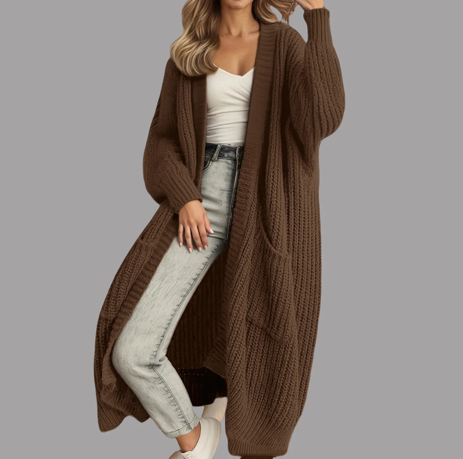 Full-length view of a women's light beige/oatmeal chunky ribbed knit duster/longline cardigan. The cardigan has a relaxed, open-front fit, dropped shoulders, and two large patch pockets. It is styled casually over a white top and blue denim jeans. Focus on the vertical rib texture and the garment's length.
