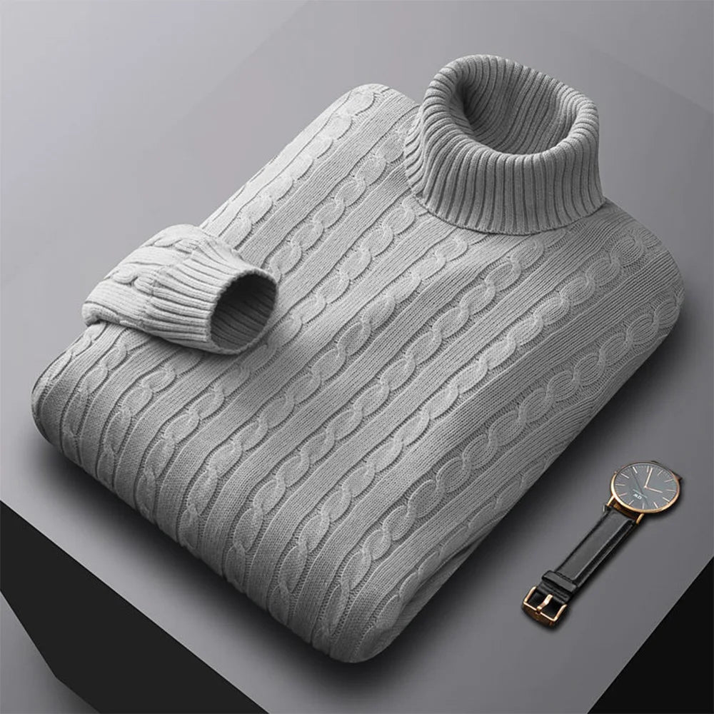 Folded men's turtleneck/roll-neck jumper in a light beige/oatmeal colour. The knit features a subtle vertical cable pattern down the body. The turtleneck and cuffs are ribbed. The sweater is styled next to a black leather men's wristwatch. Focus on the fine cable texture and the clean fold.