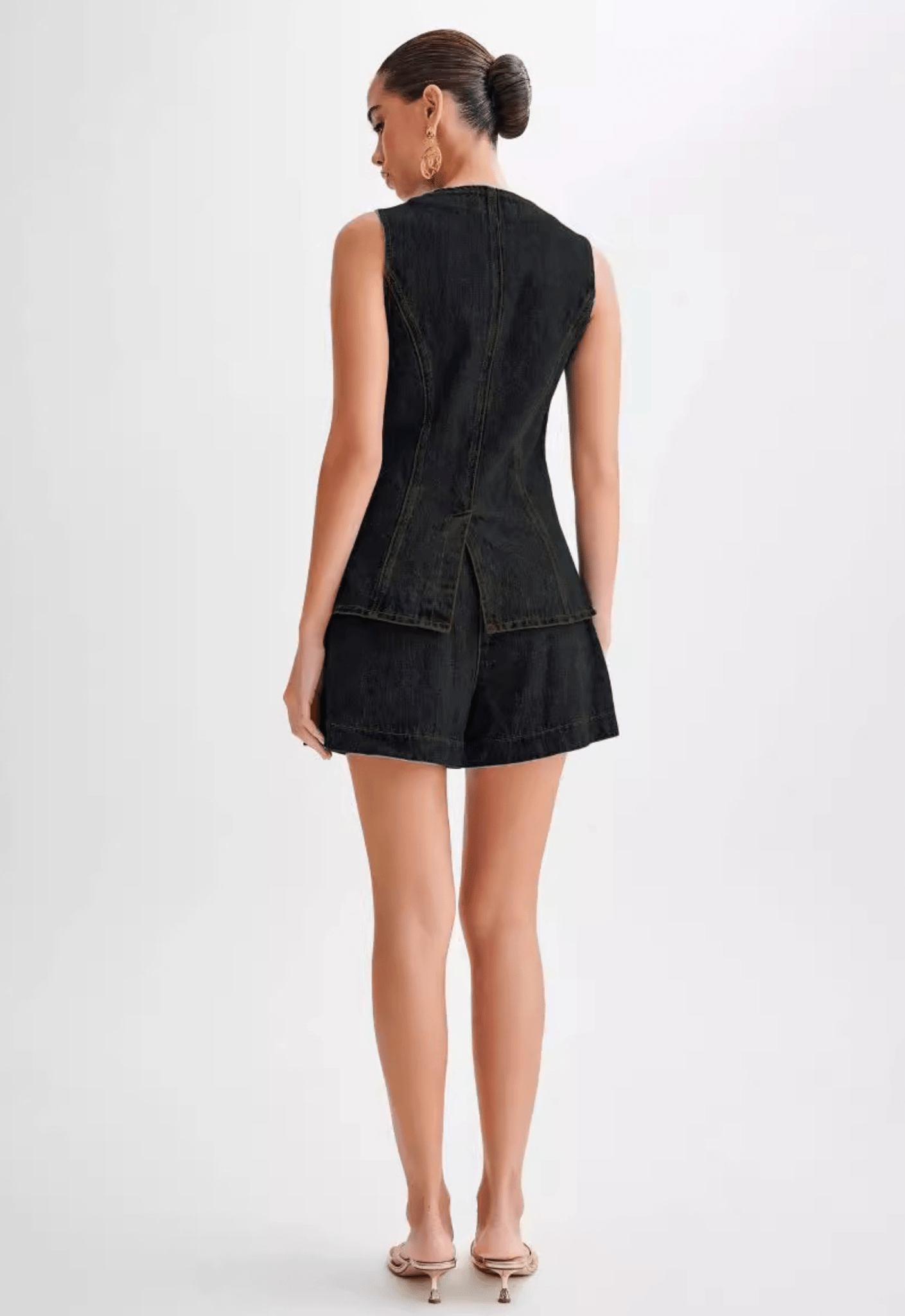 Model wearing a washed black denim sleeveless waistcoat/vest with a high crew neckline, front buttons, and a cinched waist that flares out into a structured, split peplum hem. The vest is paired with matching high-waisted denim shorts (as a set). Focus on the tailored fit and the unique peplum silhouette.