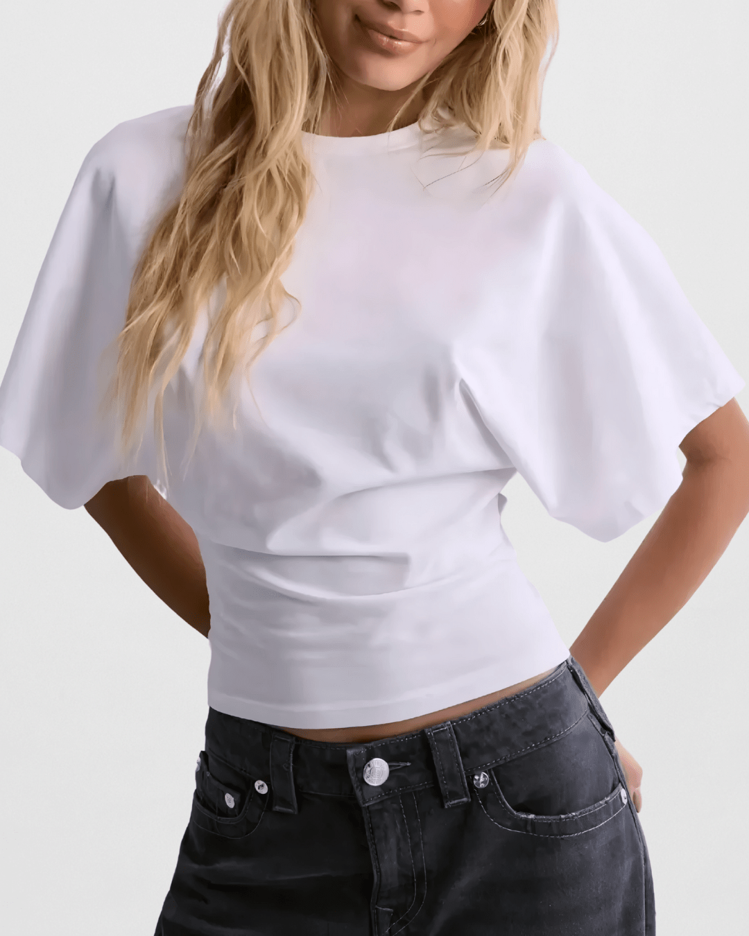 Close-up of a blonde woman wearing a white, high-neck crop top with dramatic, loose-fitting, draped dolman/batwing sleeves. The top is fitted with a cinched waistband and is styled with dark grey/washed black high-waisted denim jeans. Focus on the sharp contrast between the voluminous sleeves and the fitted hemline.