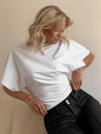 Close-up of a blonde woman wearing a white, high-neck crop top with dramatic, loose-fitting, draped dolman/batwing sleeves. The top is fitted with a cinched waistband and is styled with dark grey/washed black high-waisted denim jeans. Focus on the sharp contrast between the voluminous sleeves and the fitted hemline.