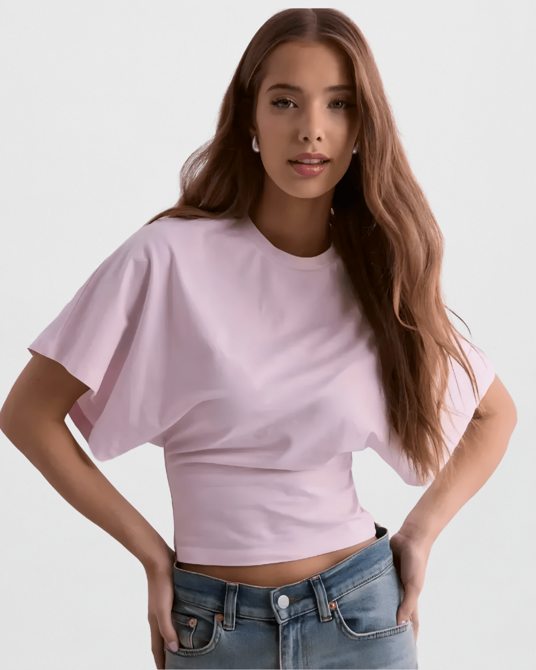 Close-up of a blonde woman wearing a white, high-neck crop top with dramatic, loose-fitting, draped dolman/batwing sleeves. The top is fitted with a cinched waistband and is styled with dark grey/washed black high-waisted denim jeans. Focus on the sharp contrast between the voluminous sleeves and the fitted hemline.