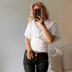 Close-up of a blonde woman wearing a white, high-neck crop top with dramatic, loose-fitting, draped dolman/batwing sleeves. The top is fitted with a cinched waistband and is styled with dark grey/washed black high-waisted denim jeans. Focus on the sharp contrast between the voluminous sleeves and the fitted hemline.