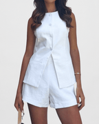 Model wearing a white, sleeveless, high-neck tailored vest made from a linen-blend fabric. The vest has a full button-up front and a distinctive long split-front hem that overlaps the matching white high-waisted linen-blend shorts (as a set). Focus on the tailored fit, the fabric texture, and the unique split-hem detail.